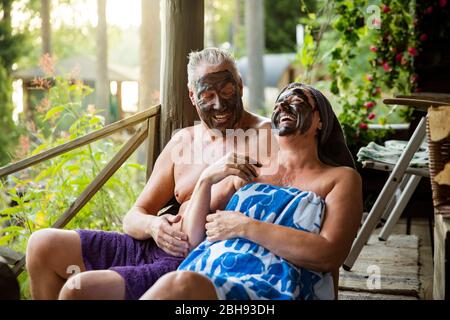 Coppia anziana con maschera facciale nera dopo sauna finlandese. Uomo maturo e donna in asciugamani seduti su gradini di legno, rilassante, ridendo e divertendosi. Foto Stock