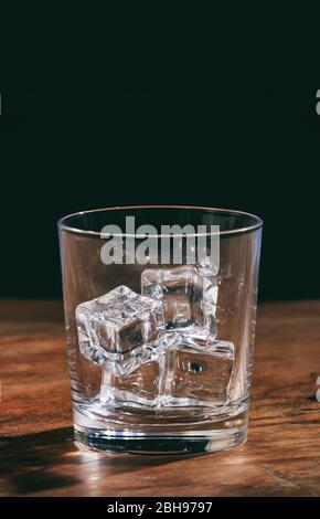 Bicchiere di whisky vuoto in cristallo con cubetti di ghiaccio sul bancone in legno. Ritratto verticale di un bicchiere trasparente trasparente trasparente trasparente pieno di rocce di ghiaccio al buio Foto Stock