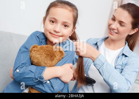 Weekend di svago. Madre spazzolatura concentrato capelli di figlia orso peloso che sorride eccitato seduta su divano primo piano Foto Stock