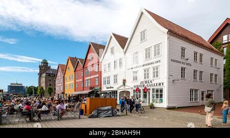 I turisti siedono ai tavoli sullo sfondo storico di case di legno colorate a Bryggen nel quartiere anseatico di Bryggen, Torget, il Molo Tedesco, Bergen, Hordaland, Norvegia, Scandinavia, Europa Foto Stock