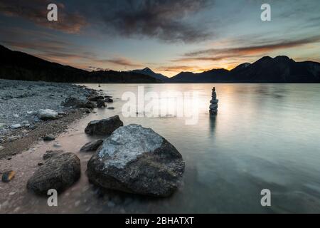 Pietre e sullo sfondo Steinmandl sulla riva di Walchensee durante il tramonto. Sullo sfondo le Prealpi bavaresi con l'Herzogstand, Heimgarten e Simetsberg. Foto Stock