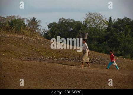 Una donna che porta un secchio di plastica sulla testa mentre cammina con i bambini su un paesaggio secco durante la stagione secca a Sumba Island, Indonesia. Foto Stock