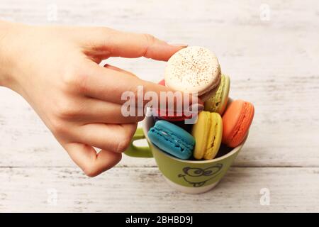 Femmina mano che prende gustosi macaroon colorati da tazza su sfondo di legno colore Foto Stock