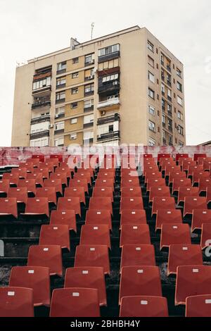 File di posti a sedere rossi vuoti nello stadio con architettura Brutalista, alto edificio di edifici di appartamenti sullo sfondo Foto Stock