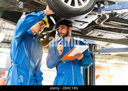 Il meccanico in uniforme blu da lavoro ispeziona il fondo dell'auto con il suo assistente. Servizio di riparazione auto, lavoro di squadra professionale. Foto Stock