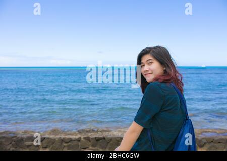 Sorridente ragazza biraciale asiatica teen seduta da sola lungo l'oceano hawaiano riposando mentre guardando l'acqua Foto Stock