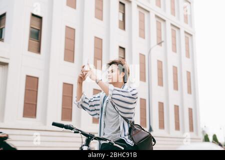 Elegante uomo chattare mentre si utilizza il telefono cellulare mentre è seduto sulla bicicletta, all'esterno. Vestito in plaid shirt, t-shirt e jeans. Close-up. Foto Stock
