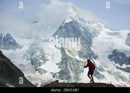 Escursionista maschile arrampicata con pali da trekking. Incredibile montagna rocciosa sulla neve nelle Alpi Pennine sullo sfondo. Escursioni in montagna, uomo che raggiunge il picco. Natura selvaggia con viste mozzafiato. Turismo sportivo nelle Alpi. Foto Stock