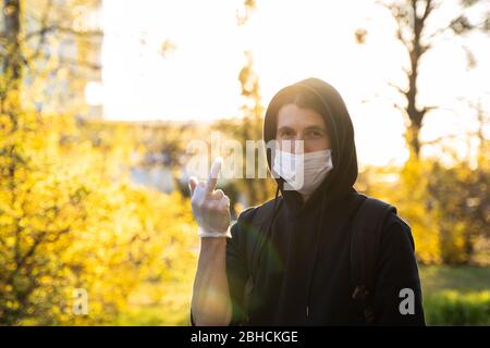 Uomo con felpa con cappuccio nera che indossa una maschera medica con un dito medio. Prevenzione di infezione batterica Coronavirus o epidemia di Covid 19 su una strada Foto Stock