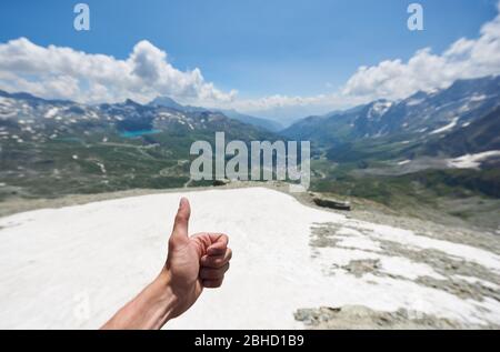 Primo piano della mano dell'uomo facendo gesto di approvazione con colline erbose e cielo bello sullo sfondo. Alpinista che mostra i pollici fino segno mentre escursioni in montagna. Concetto di viaggio e alpinismo. Foto Stock