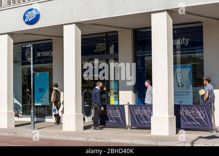 Persone coda fuori dalla farmacia Boots durante il blocco Coronavirus 19 a Canterbury, Kent UK, UN uomo di sicurezza indossa una maschera facciale. Foto Stock