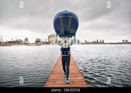 Atleta muta in porta il suo paddleboard al molo in legno presso il lago a freddo contro il cielo nuvoloso Foto Stock