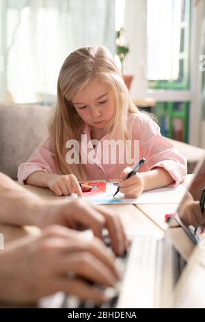 Adorabile bambina con lunghi capelli biondi disegno con evidenziatori mentre seduto da tavolo accanto al padre che naviga in rete Foto Stock