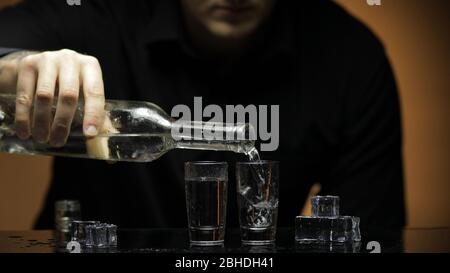 Barman che versa la vodka congelata da una bottiglia in due bicchieri da bicchierini con cubetti di ghiaccio su sfondo nero. Barman versare in bicchiere di vetro espresso una bevanda alcolica trasparente trasparente trasparente al rum tequila Foto Stock