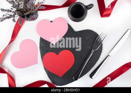 San Valentino amore cena romantica messa a punto su sfondo bianco, nastro rosso, piatto a forma di cuore nero e fiori Foto Stock