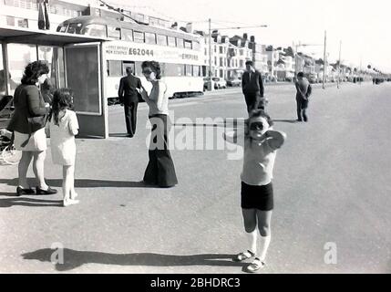 Vita quotidiana per le strade di Blackpool, Inghilterra, nel 1974. Una giovane ragazza sul lungomare di Blackpool, Lancashire, Regno Unito, copre le orecchie con le mani per ignorare la sua famiglia che sta parlando nelle vicinanze. Un tram passa sullo sfondo. Foto Stock