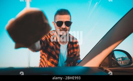 Giovane uomo pulendo con cura un parabrezza sulla sua auto con un panno in microfibra. Foto Stock