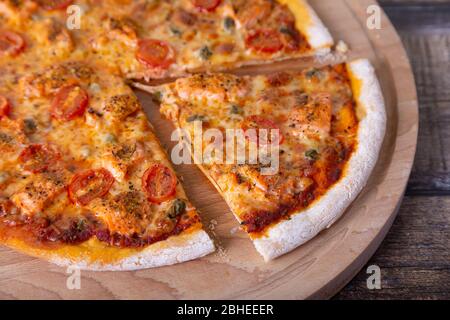 Pizza con salmone, pomodori e capperi su un asse di legno. Pizza intera, un pezzo tagliato. Primo piano. Foto Stock