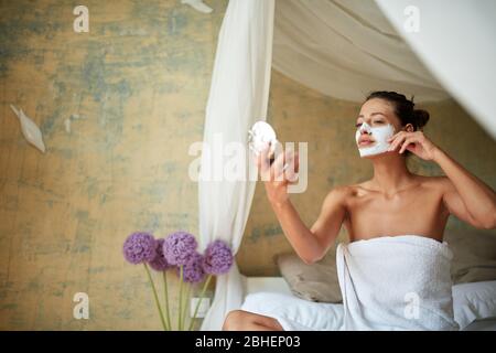 Giovane ragazza allegra maschera facciale sul viso seduta a letto e guardando specchio.bellezza e il concetto di cura del corpo. Foto Stock