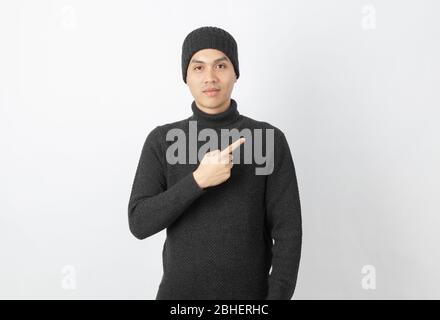 Giovane uomo asiatico bello che indossa maglione grigio e beanie che punta al lato con le dita per presentare un prodotto o un'idea su sfondo bianco. Foto Stock