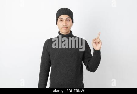 Giovane uomo asiatico bello che indossa maglione grigio e beanie che punta al lato con le dita per presentare un prodotto o un'idea su sfondo bianco. Foto Stock