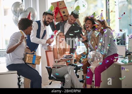 colleghi che sorprendono la collega femmina con torta di compleanno al lavoro in ufficio. sorpresa, festa di compleanno al lavoro, affari informali Foto Stock