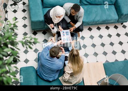 Vista dall'alto di quattro giovani imprenditori di diverse nazionalità che lavorano con documenti finanziari e guardano insieme grafici e grafici, seduti Foto Stock