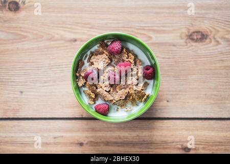 Vista dall'alto di un pasto sano di cereali integrali. Colazione a base di latte, lamponi e cereali. Ciotola verde su sfondo tavolo da cucina in legno Foto Stock