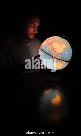 Un uomo che guarda un globo rotante Terra lampada luce illuminata scrivania Mappa Geografia nella notte buia con il volto illuminato Foto Stock