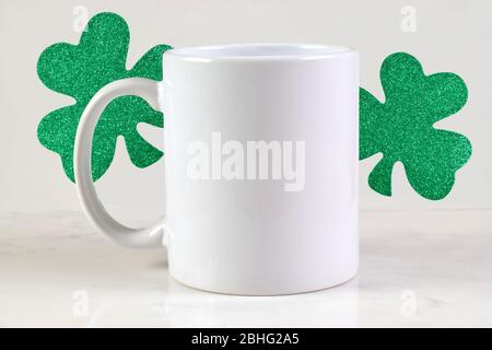 Tazza da caffè bianca da 11 once, seduta felicemente su uno sfondo di marmo bianco circondato da scintillanti sciamrocks del giorno di San Patrizio. Foto Stock
