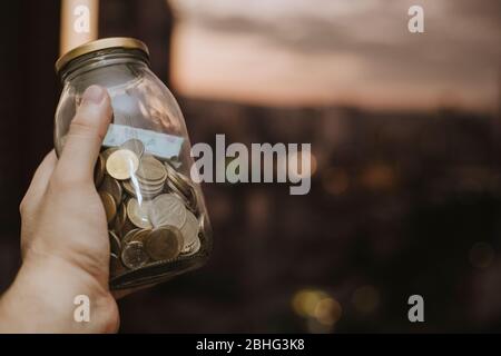 Uomo ispanico che tiene a mano un vaso di risparmio con banconote e monete in valuta colombiana in una serata tramonto Foto Stock