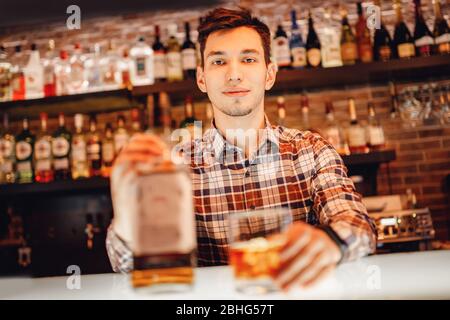 Il barman versa la frusta dalla bottiglia nel bicchiere di ghiaccio bar. Sfondo marrone scuro Foto Stock
