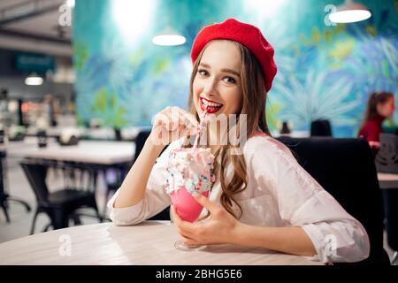 Francia dolce dessert nazionale, ragazza in beretto rosso mangia torta rosa, caramella floss e milkshake, ride e sorrisi Foto Stock