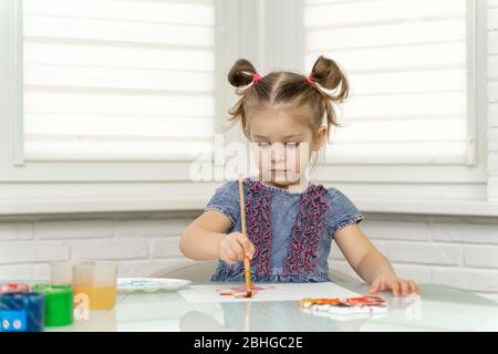 una bambina in un abito blu dipinge alla finestra, ha preso un pennello e si muove su carta. rimanere a casa, virus covid-19, auto-isolamento, isolamento Foto Stock