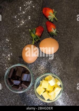 Due ciotole di vetro riempite con cioccolato e burro su un piano scuro, due uova e alcune fragole Foto Stock