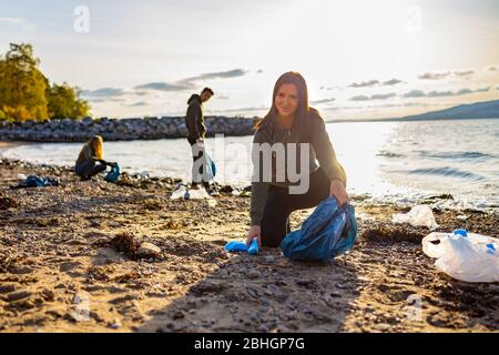 Sorridente giovane donna che pulisce la spiaggia con volontari durante il tramonto Foto Stock