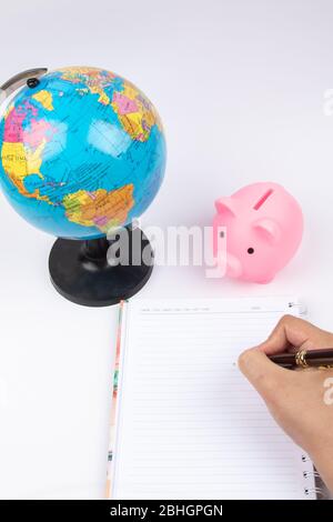 Penna per mano da donna con notebook vuoto aperto con globo terrestre e piggy bank sulla scrivania dell'ufficio. Foto Stock