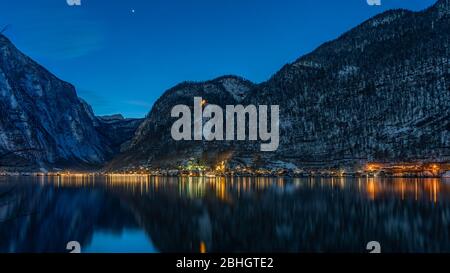 Villaggio austriaco innevato Hallstatt dal lago circondato da montagne innevate con luci accese dopo il tramonto di notte Foto Stock