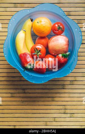 Ciotola blu con frutta fresca e pulita su una base di legno. Banane, pomodori, mele, fragole e arance. Foto Stock