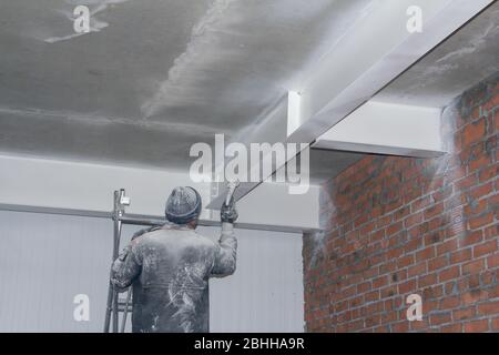 Un lavoratore dipinge un soffitto da una pistola a spruzzo. Lavori di verniciatura in un cantiere. Foto Stock