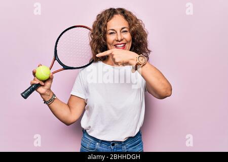 Media età bella sportswoman giocare a tennis tenendo racchetta e palla su sfondo bianco sorridendo felice puntando con mano e dito Foto Stock