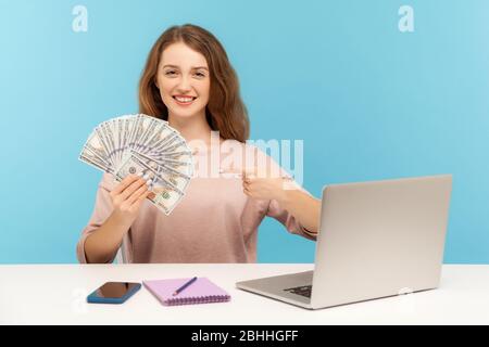 Scommesse online, guadagni su internet. Donna positiva che punta ai soldi, freelancer che ci mostra le fatture del dollaro, seduta al posto di lavoro con i offi domestici del laptop Foto Stock