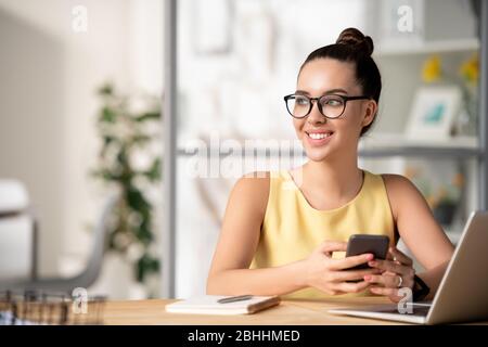 Responsabile creativo ispirato pensivo negli occhiali seduti alla scrivania con computer portatile e telefono per il controllo in ufficio Foto Stock