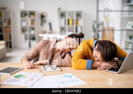 Giovani colleghi esausti appoggiati al tavolo con documenti di report e notebook mentre dormono sul posto di lavoro Foto Stock