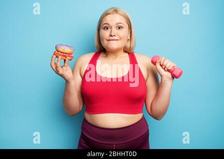 Fat ragazza pensa di mangiare ciambelle invece di fare palestra. Concetto di indecisione e dubbio Foto Stock