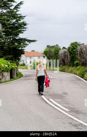 Una donna bionda cammina lungo una strada verde ai lati e case sullo sfondo. Vista posteriore. Indossa jeans neri, una maglietta bianca e in mano un Foto Stock