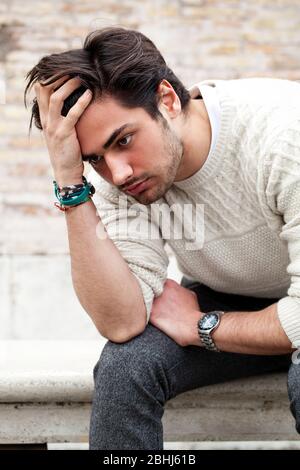 Giovane uomo preoccupato seduto e fissando all'aperto con le mani sulla testa e capelli con un atteggiamento disperato. Maglione bianco. Per strada. Foto Stock