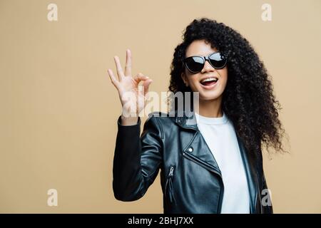 La donna alla moda dai capelli ricci dice che tutto è bello, fa un gesto giusto, dice sì alle nuove opportunità, indossa occhiali da sole alla moda e giacca in pelle nera, Foto Stock