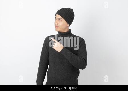 Giovane uomo asiatico bello che indossa maglione grigio e beanie che punta al lato con le dita per presentare un prodotto o un'idea su sfondo bianco. Foto Stock