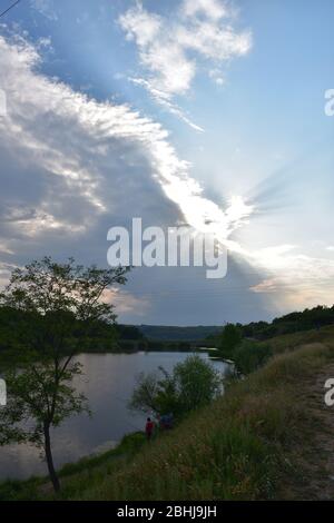 Accanto al lago, due persone pescano nel verde del lago. Nel cielo, il sole si rompe dietro le nuvole Foto Stock